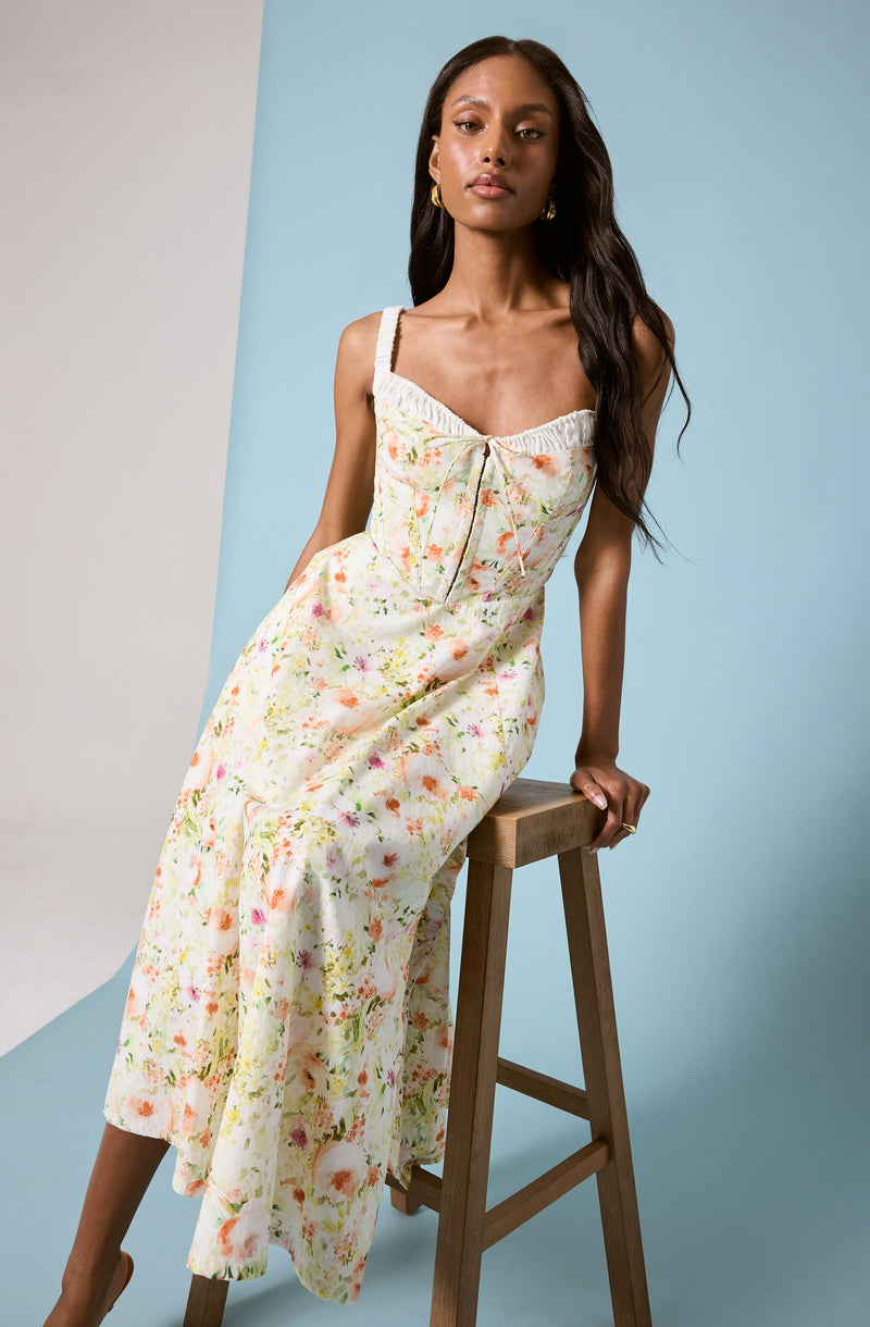 A woman is wearing a sleeveless, light-colored floral dress with a fitted bodice that has a gathered neckline and a bow tie detail at the front. The dress features wide shoulder straps and a flowing, ankle-length skirt with a soft, pastel floral print. She has long, dark, wavy hair, and is accessorized with gold hoop earrings. She is seated on a wooden stool against a simple background with soft neutral and blue tones.