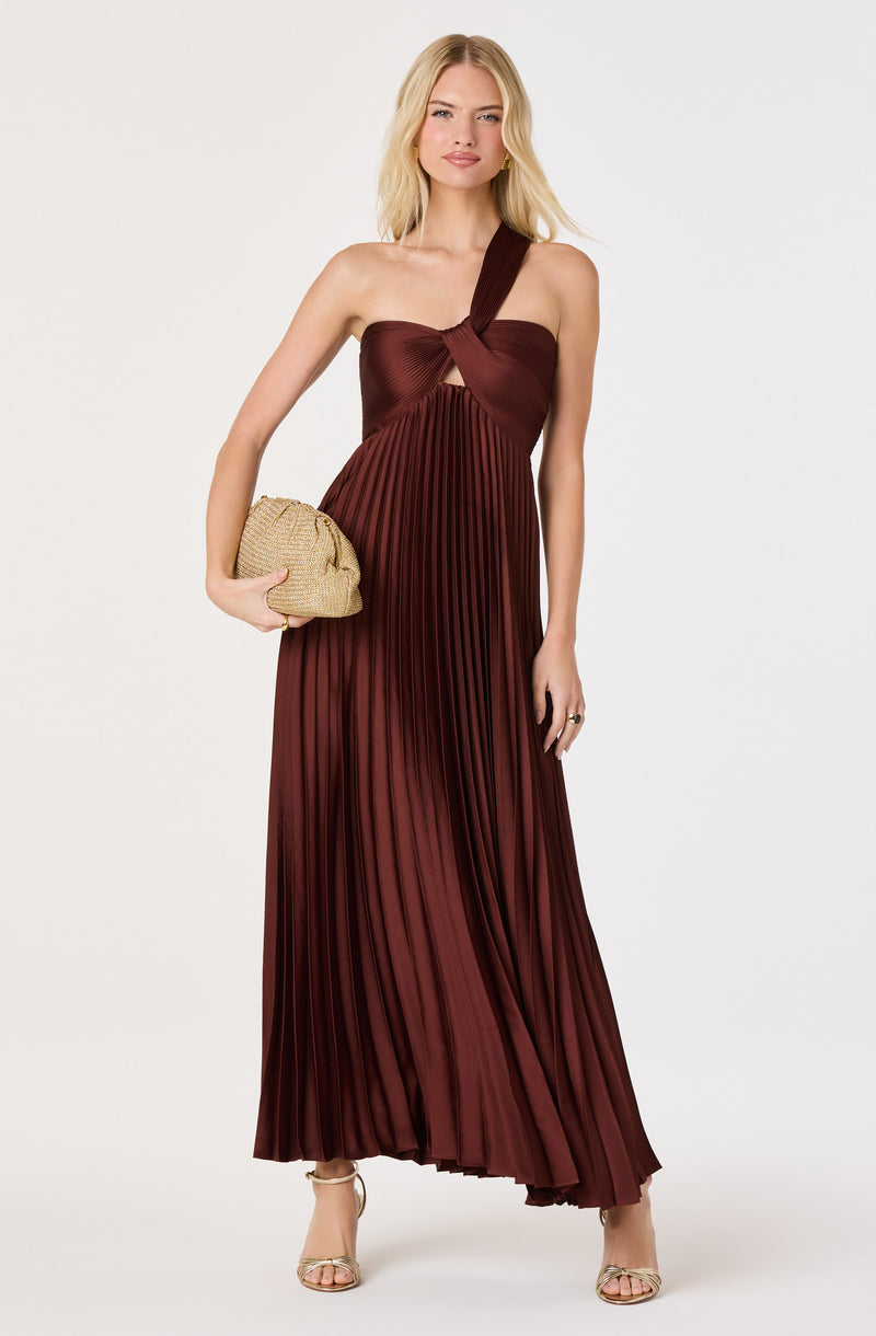 Woman in a burgundy pleated evening gown with a one-shoulder strap and knot detail, holding a beige woven clutch.
