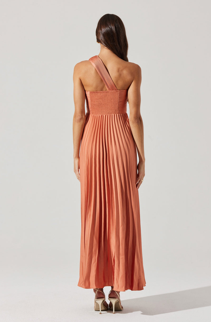 A woman is wearing a floor-length, pleated dress in a warm, peachy-orange color. The dress has an asymmetrical strap over her left shoulder, with the bodice fitted and textured, and the skirt featuring wide, vertical pleats. She is standing with her back to the camera, her long brown hair falling down her back.