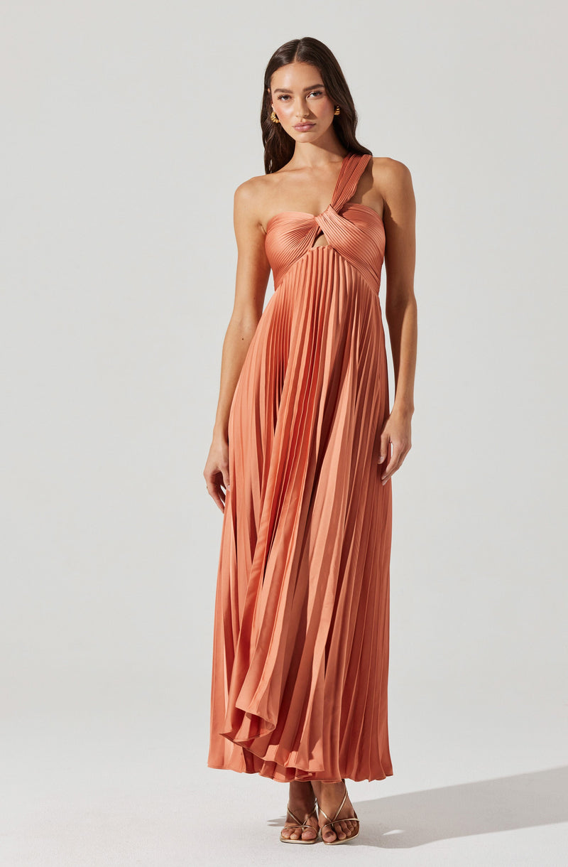 A woman is wearing a floor-length, pleated, rust-colored dress with a one-shoulder design. The dress features a twisted knot detail at the bust and has an asymmetrical strap on one shoulder, while the other shoulder is bare. The fabric appears shiny, adding a smooth texture to the pleats. She is also wearing open-toe high-heeled sandals.