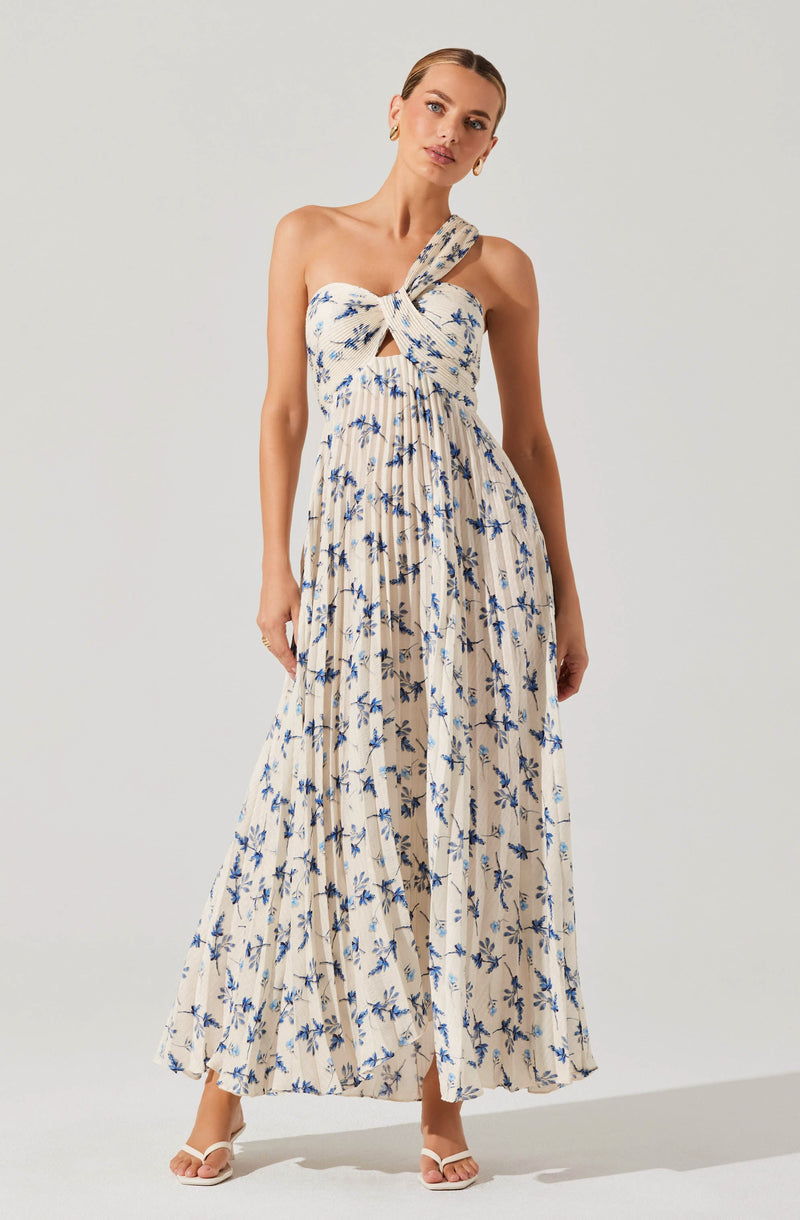 A woman is wearing a sleeveless, long maxi dress with a light beige background and a blue floral print. The dress has a distinctive twisted knot detail at the bust with a small keyhole opening just below it. The skirt is flowing and pleated, creating a soft, airy silhouette.