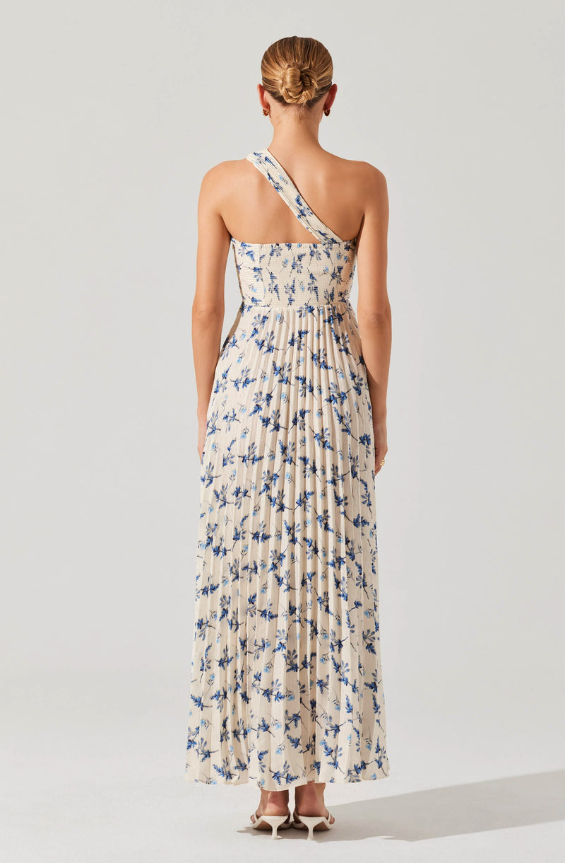 A woman is wearing a long, flowing dress with a beige base color and a blue floral print. The dress features a one-shoulder strap design. The fabric appears lightweight and comfortable, suitable for warm weather. She is also wearing neutral-colored high heels.