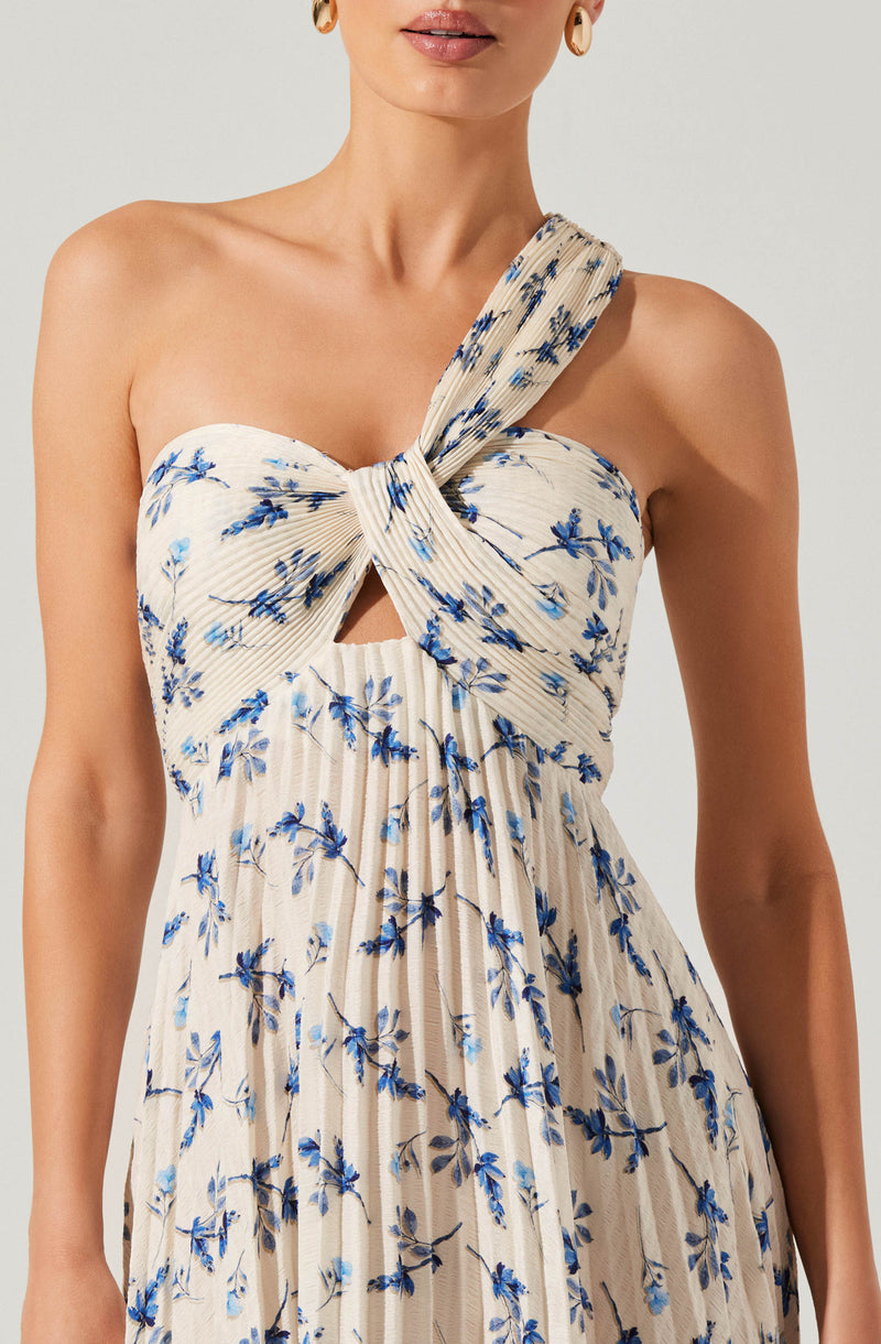 A woman is wearing a strapless dress with a knot and bow detail at the top. The dress features a cream-colored background with a blue floral print, including small leaves and branches. The fabric has vertical pleats, and a small cutout is present just below the knot. One shoulder has an additional fabric strap that extends from the knot.