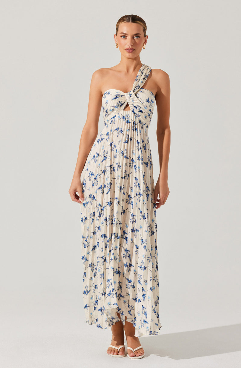 A woman is wearing a sleeveless, strapless maxi dress with a large bow at the front. The dress features a beige base color with a blue floral pattern, and it has a cut-out detail just below the bow. The fabric is lightweight and pleated, flowing down to her ankles. She is also wearing white heeled sandals.