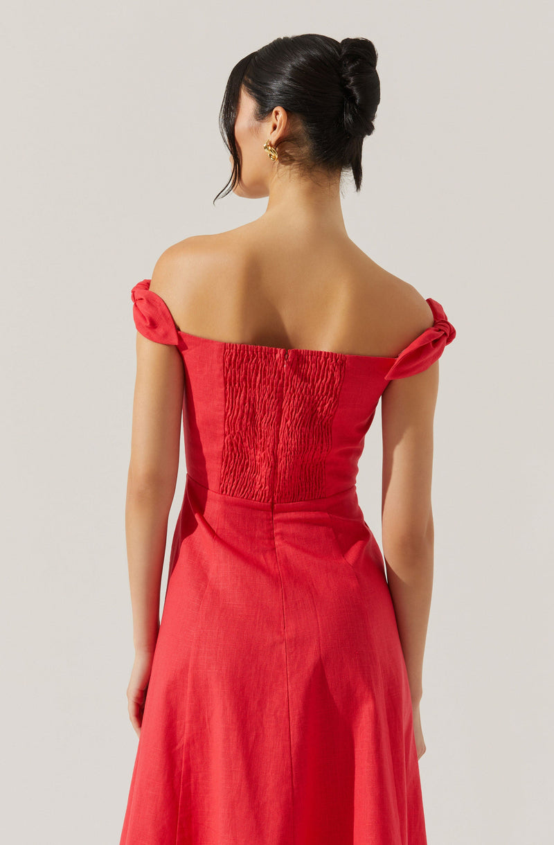A woman is wearing a red off-the-shoulder dress with gathered fabric details on the bodice and short, tied sleeves. The dress has a fitted waist and a long, flowing skirt.
