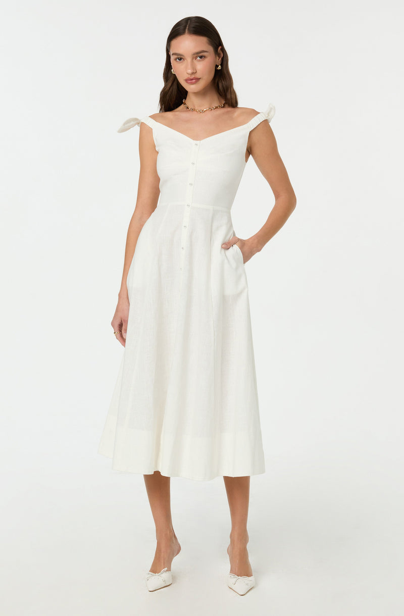 A woman is wearing a white, sleeveless, button-front dress with a fitted bodice and a full, mid-length skirt. The dress features off-the-shoulder straps tied into bows on her shoulders. She is also wearing white pointed-toe shoes with a bow detail on the front.