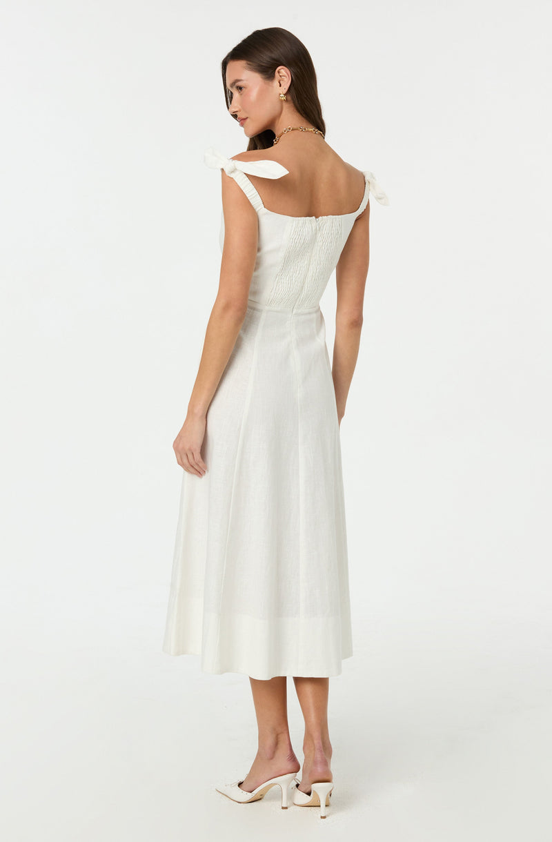 A woman is wearing a sleeveless, white midi dress with shoulder ties that form bows. The dress features a fitted, shirred bodice and a flowy, A-line skirt that reaches just above her ankles. She is also wearing white high-heeled shoes with a pointed toe and slim heel.