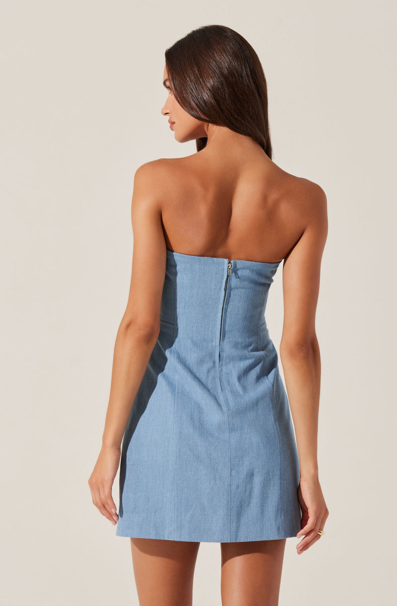 A woman is wearing a strapless, light blue denim dress that is fitted at the top and flares out slightly at the bottom. The dress features a visible metal zipper running down the center of the back.
