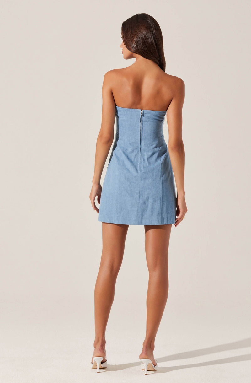 A woman is wearing a strapless, light blue denim mini dress with a visible back zipper closure. She has nude high heels and long, dark brown hair.