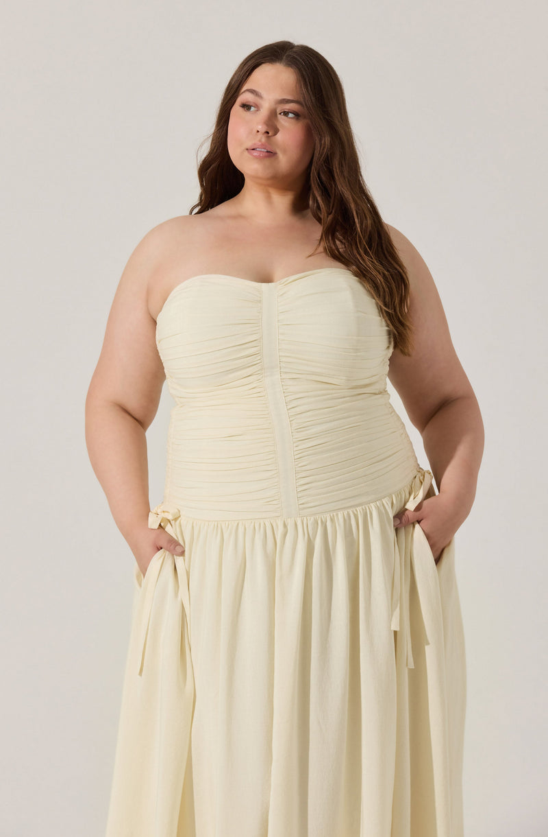 A woman wearing a strapless cream-colored dress with a ruched bodice, a front zipper, and a gathered waist with ties. The skirt is full and flowing, made of a lightweight fabric.