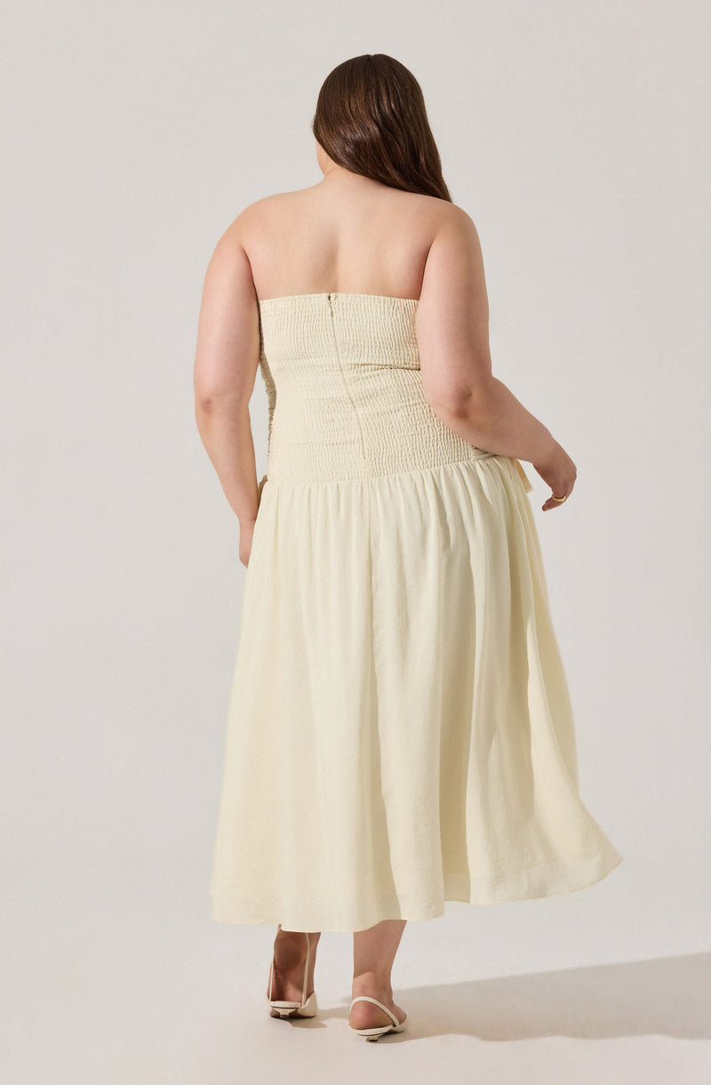 A woman is wearing a strapless, cream-colored dress with a smocked bodice and a flowing, gathered skirt. She has long, brown hair and is barefoot, standing against a plain, light-colored background.