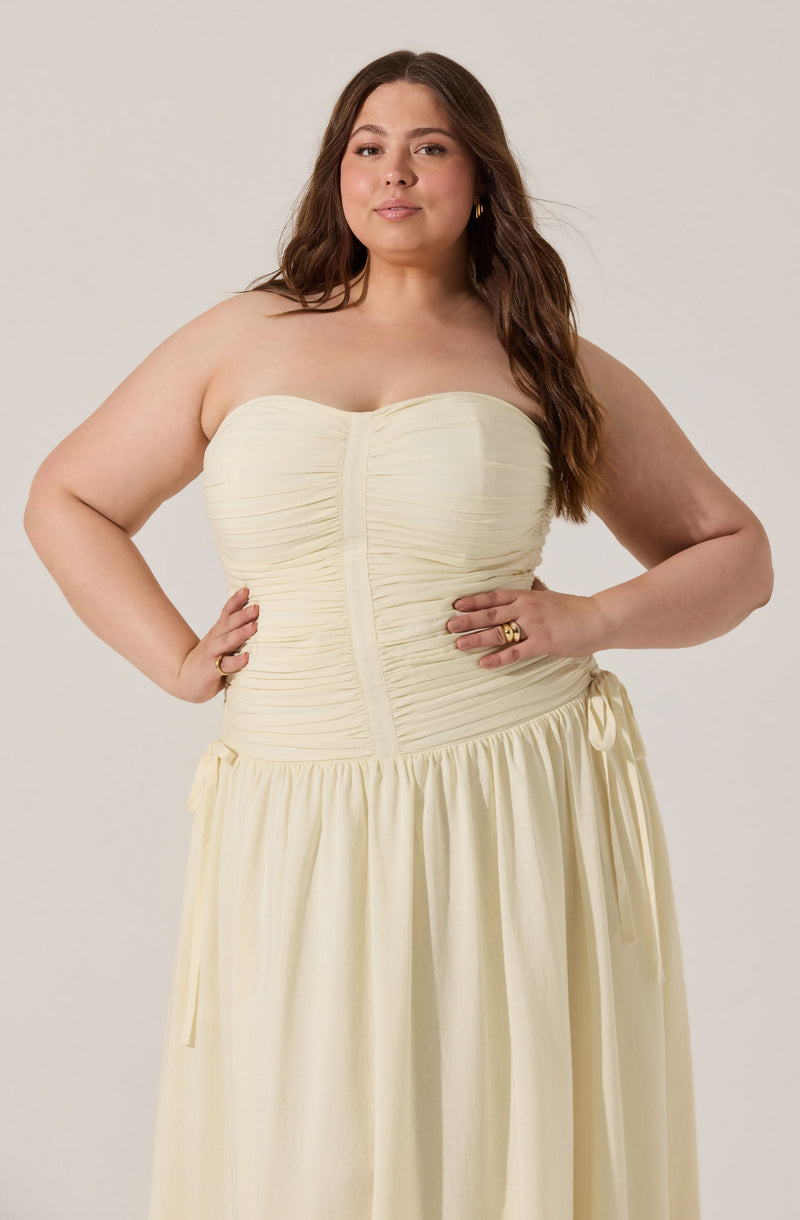 A woman is wearing a cream-colored, strapless dress with a ruched bodice and a gathered skirt. The dress features a front seam down the center of the bodice and has side ties at the waist.