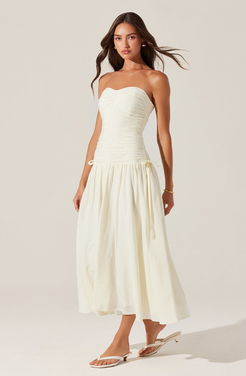 A woman is wearing a strapless white dress with a ruched bodice that fits snugly around her torso, and a full, flowing skirt that reaches mid-calf. She has on white, open-toe heeled sandals and is accessorized with small hoop earrings and a gold bracelet. Her long, dark hair is styled in loose waves.