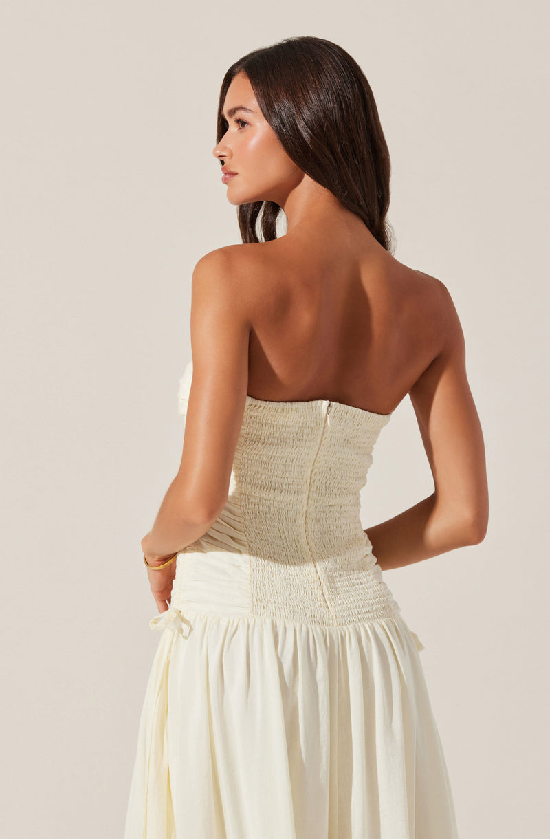 A woman is wearing a strapless, cream-colored, textured dress with a fitted bodice and a gathered, flowing skirt. The dress has a side zipper and a small bow detail on the side of the skirt.