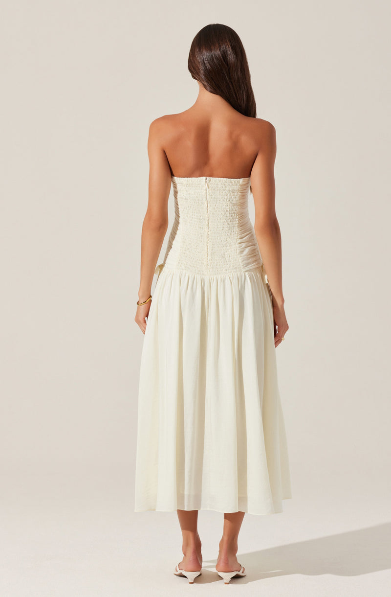 A woman is wearing a strapless, off-white, smocked dress with a fitted bodice that extends down to the waist. The skirt is long, flowing, and gathered, reaching above her ankles. She has long, dark hair and is wearing a gold bracelet on her left wrist.