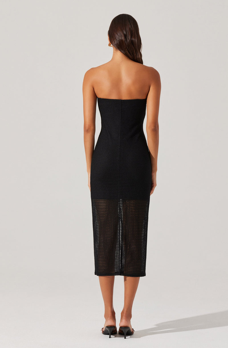 A woman is wearing a black strapless dress that extends from her chest to mid-calf. The dress has a textured, perforated pattern with small holes, which become more visible below the waistline. She is also wearing black open-toe high heels. Her long, dark brown hair is worn down and straight.