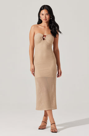 A woman is wearing a strapless, beige, textured knit dress that reaches mid-calf. The dress features a gathered detail at the bust with a small decorative element in the center. She is also wearing black, strappy high-heeled sandals.