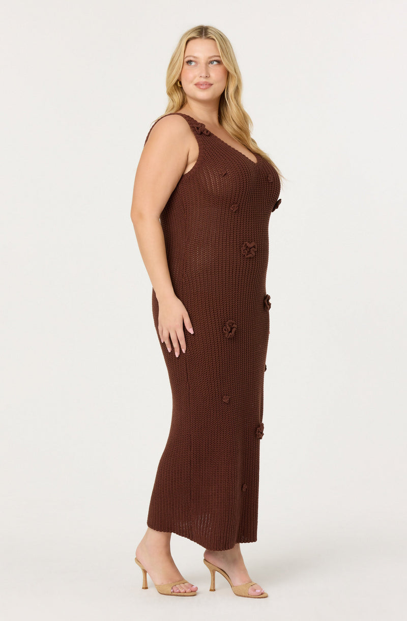 A woman is wearing a sleeveless, ankle-length dress made of a chunky, dark brown crochet material. The dress features decorative crochet flowers along the front and also has ruffle details on the shoulder straps. She is wearing nude, open-toe high heels.