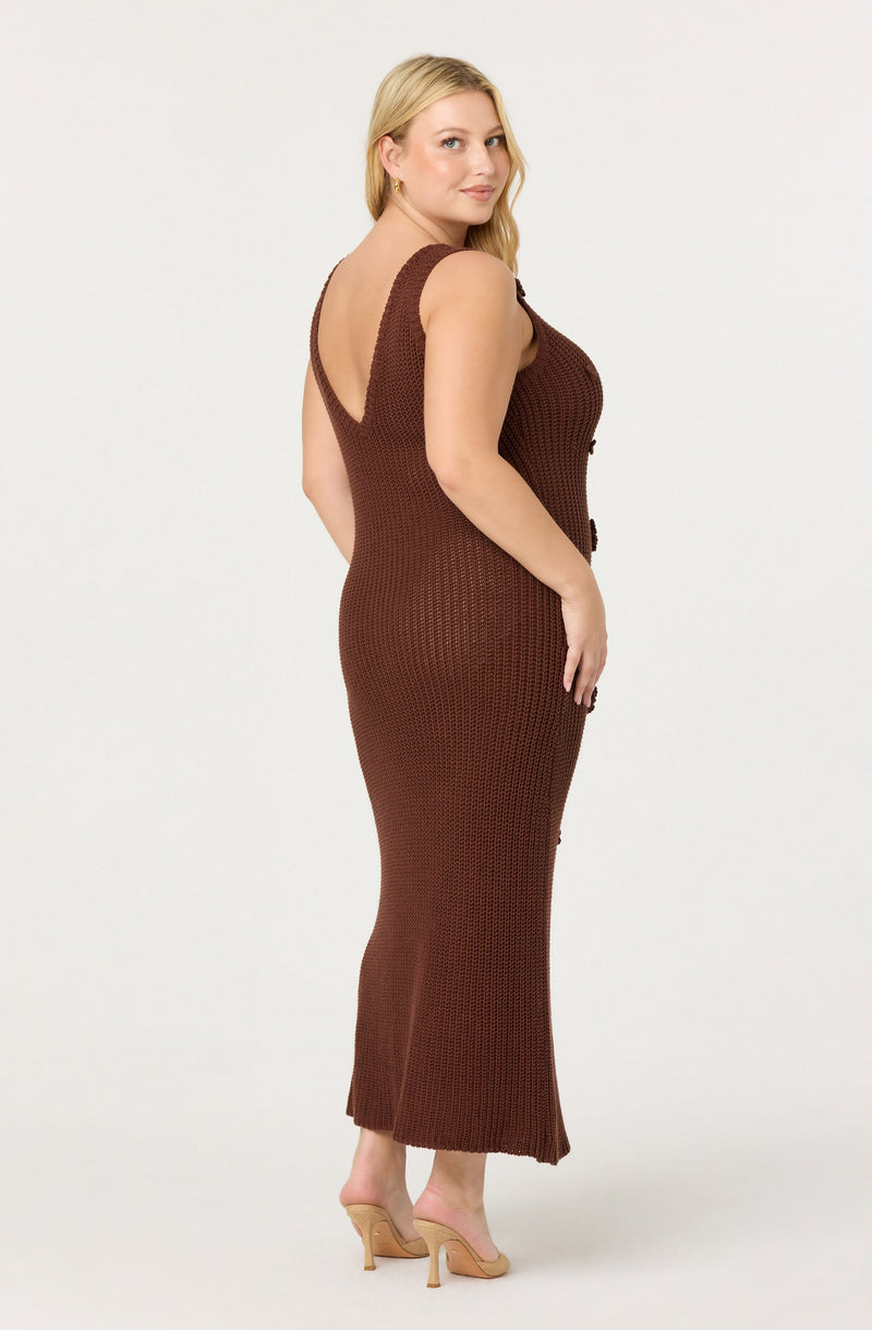 A woman is wearing a sleeveless, deep V-neck, brown knitted maxi dress with a fitted silhouette. She is also wearing beige high-heeled shoes.