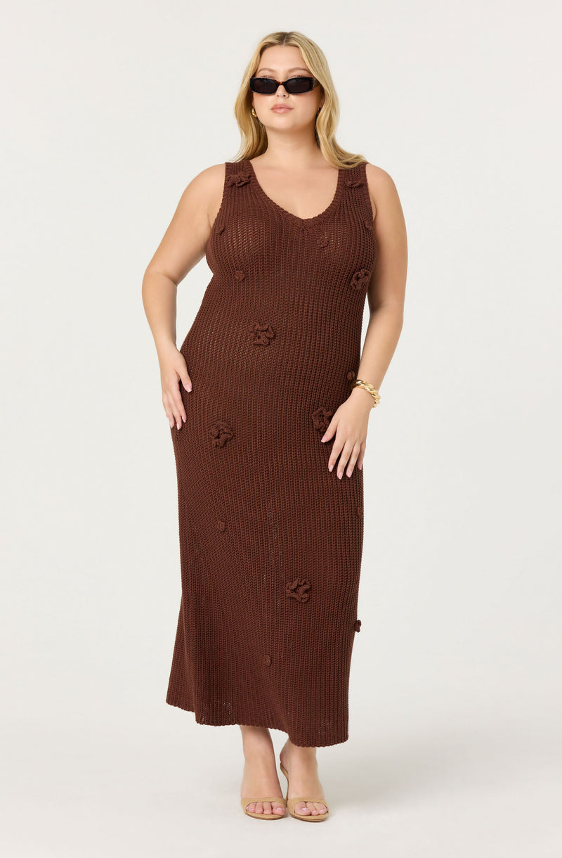 A woman is wearing a sleeveless, long brown crochet dress with a V-neckline. The dress features some open-knit patterns and decorative knot details scattered throughout. She pairs the outfit with nude open-toe heels, dark sunglasses, and a gold bracelet.