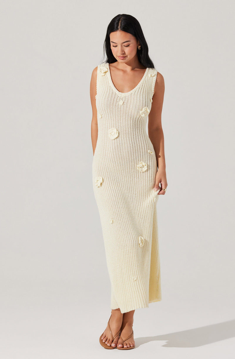 A woman is wearing a sleeveless, cream-colored, knit maxi dress with a scoop neckline. The dress features decorative flower appliqués scattered across the fabric. She is also wearing clear, open-toe high heels.