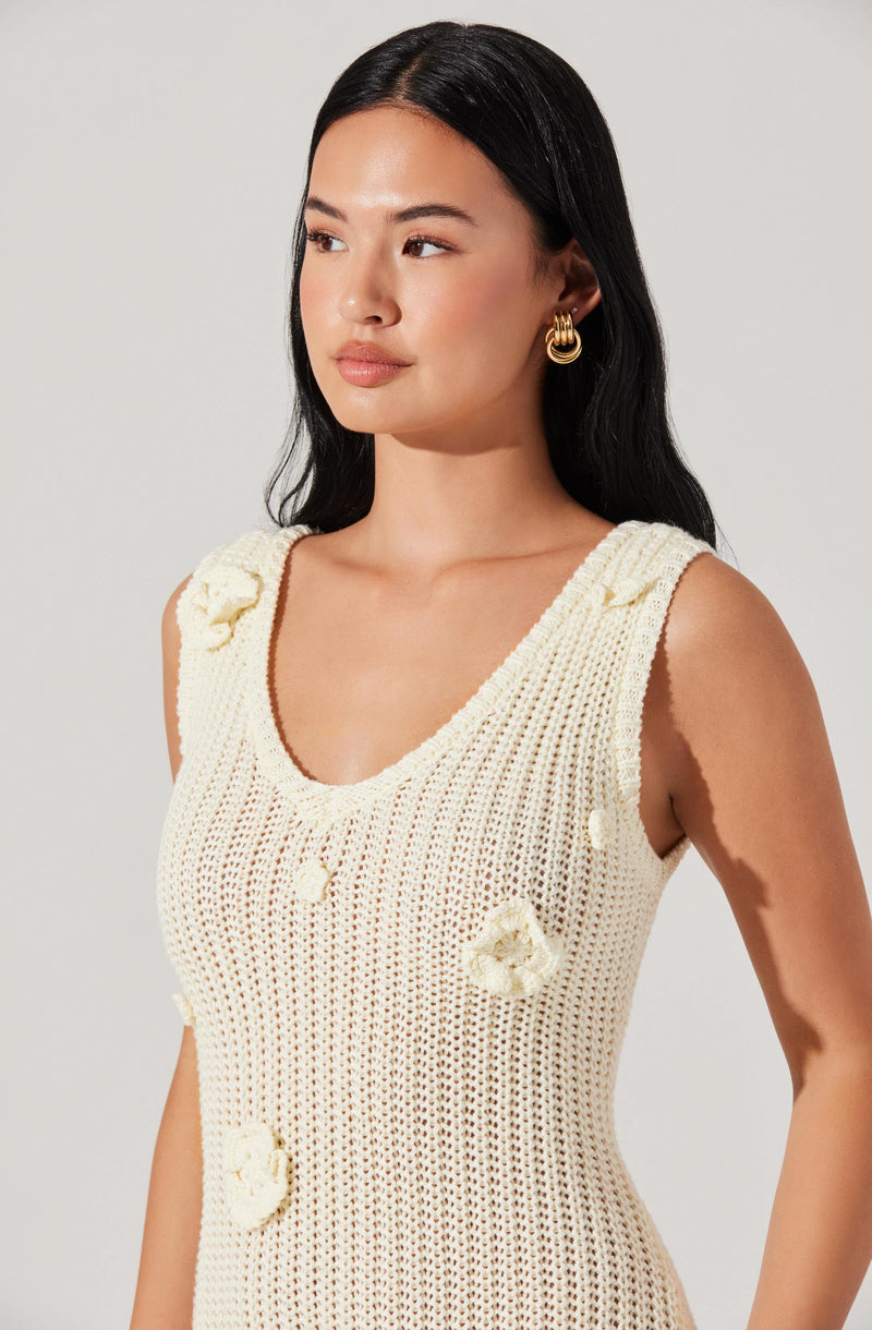 A woman wearing a sleeveless, cream-colored, open-knit crochet dress with floral adornments. The dress features embroidered flower details on the shoulders and chest areas.
