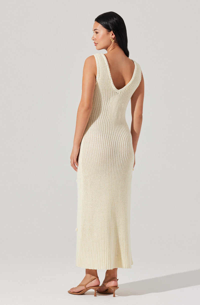 A woman is wearing a cream-colored, sleeveless, knit maxi dress with a deep V back. The dress features a textured, open-knit pattern throughout. She is also wearing beige high-heeled sandals and gold hoop earrings.