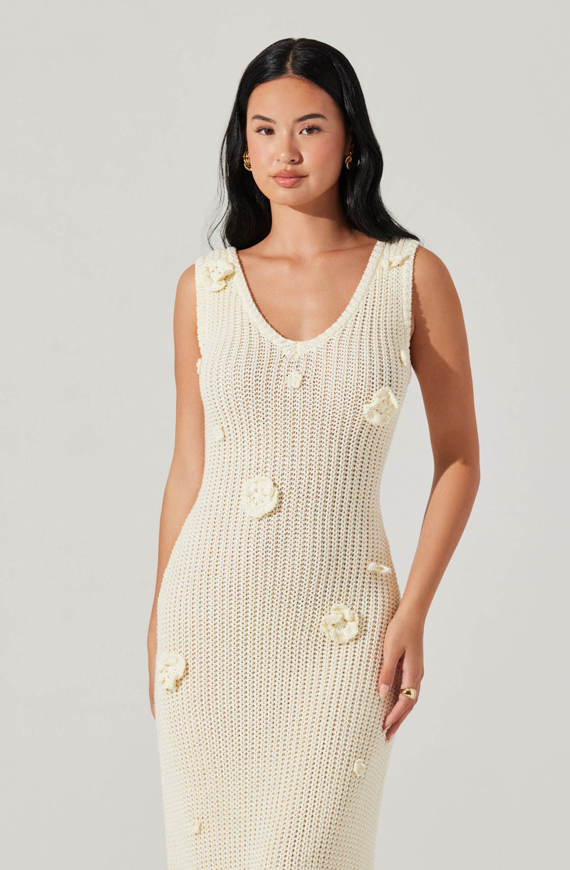 A woman is wearing a sleeveless, cream-colored knit dress with a scoop neckline. The dress features decorative fabric flowers sewn onto it, adding texture and visual interest. She has long, dark hair, gold earrings, and a gold ring on her right hand.