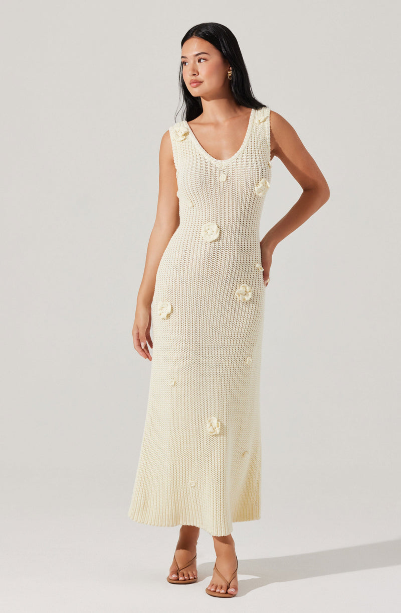 A woman is wearing a sleeveless, cream-colored, knit dress that extends to her ankles. The dress features a scooped neckline and is decorated with textured floral appliqués scattered throughout. She is also wearing transparent-strap nude heels.