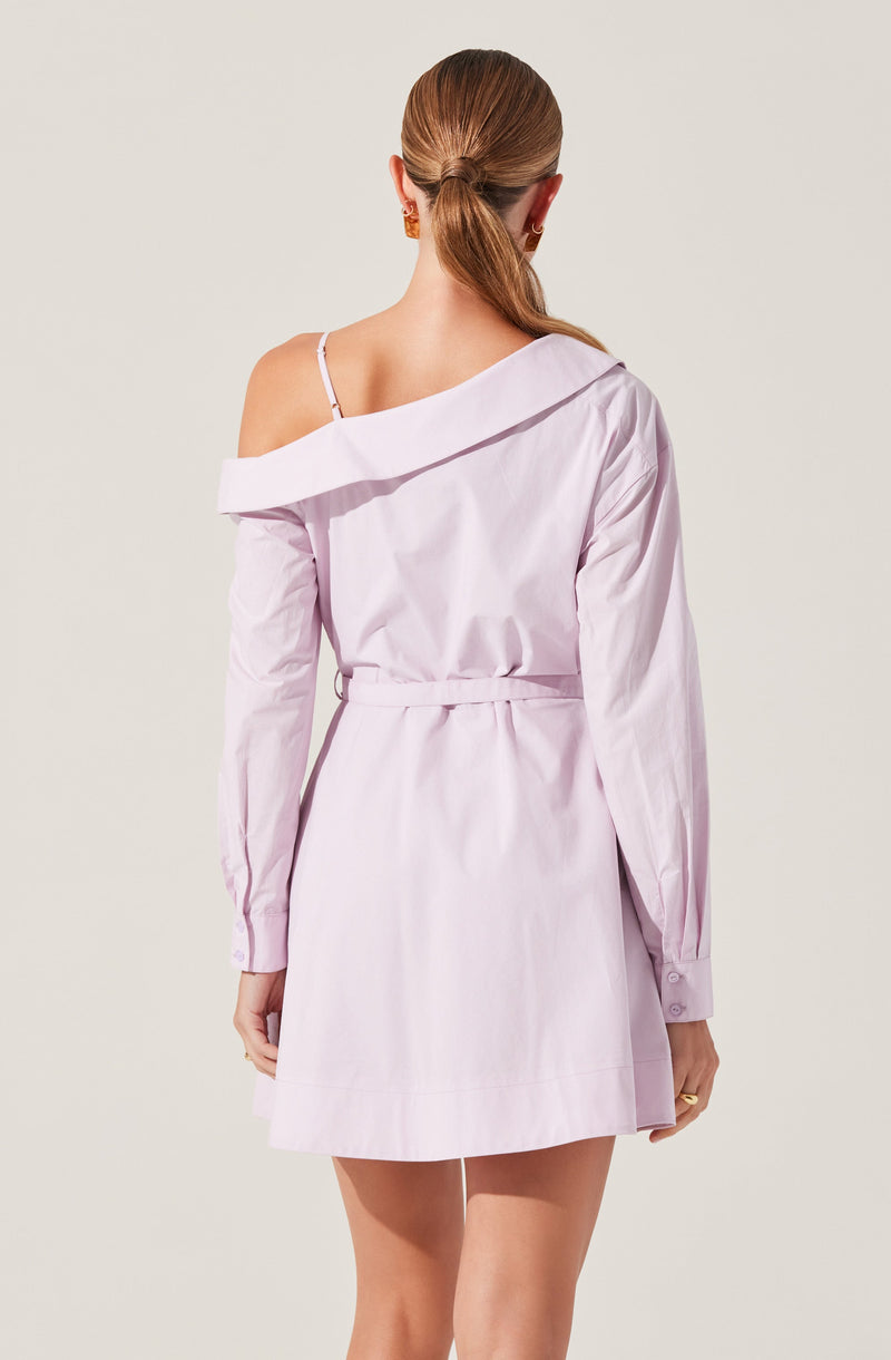 A woman is wearing a light pink shirt dress with long sleeves and buttoned cuffs. The dress has an off-the-shoulder design, exposing one shoulder with a strap, and features a collar that falls off one side. The dress is belted at the waist, creating a cinched look, and has a short hemline above the knees.