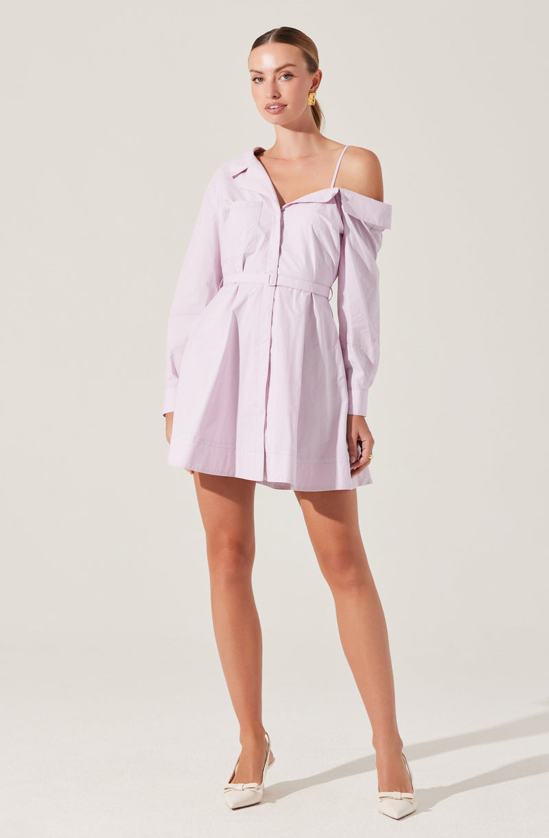 A woman is wearing a light pink, off-the-shoulder shirt dress with a collar and long sleeves. The dress has a gathered waist, button details, and a slightly flared skirt. She is also wearing a white spaghetti strap top underneath, with pointed white heeled shoes.