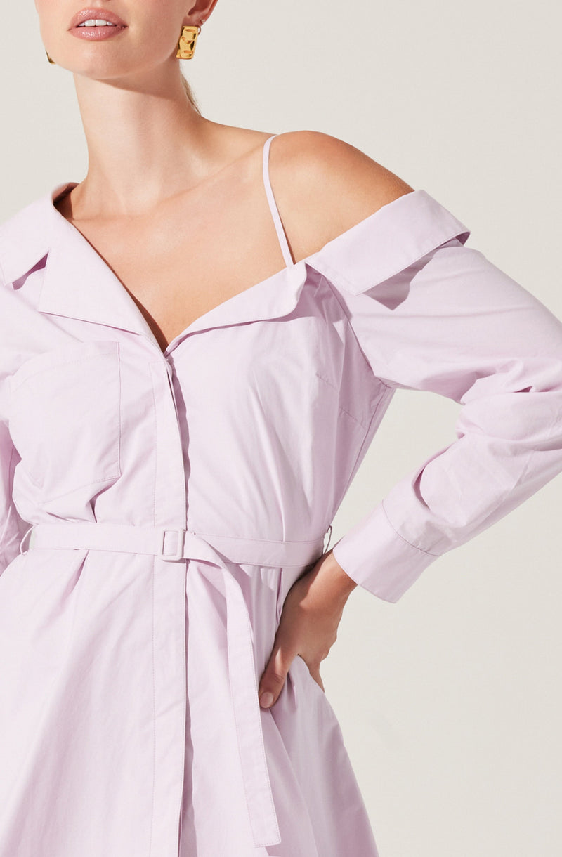 A woman is wearing a light pink, off-the-shoulder shirt dress with long sleeves, featuring a collar and a waist tie. She has a white strap visible on her shoulder, and she is accessorized with a gold earring.