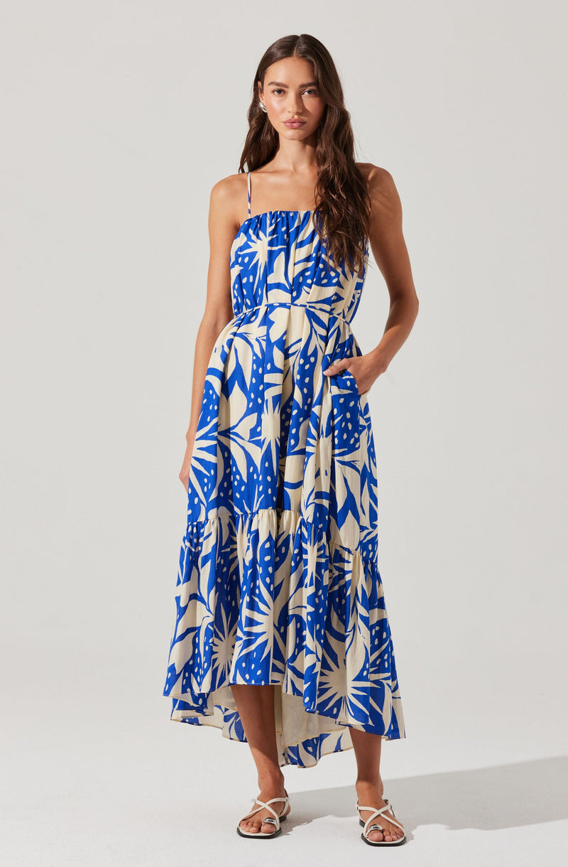 A woman is wearing a sleeveless, ankle-length dress with thin spaghetti straps. The dress features a bold, abstract tropical print in shades of blue and cream. It has a loose, flowing fit with a gathered bodice and a waistband that ties at the side. She is also wearing open-toe, strappy sandals in a light color.