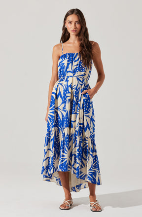 A woman is wearing a sleeveless, ankle-length dress with thin spaghetti straps. The dress features a bold, abstract tropical print in shades of blue and cream. It has a loose, flowing fit with a gathered bodice and a waistband that ties at the side. She is also wearing open-toe, strappy sandals in a light color.
