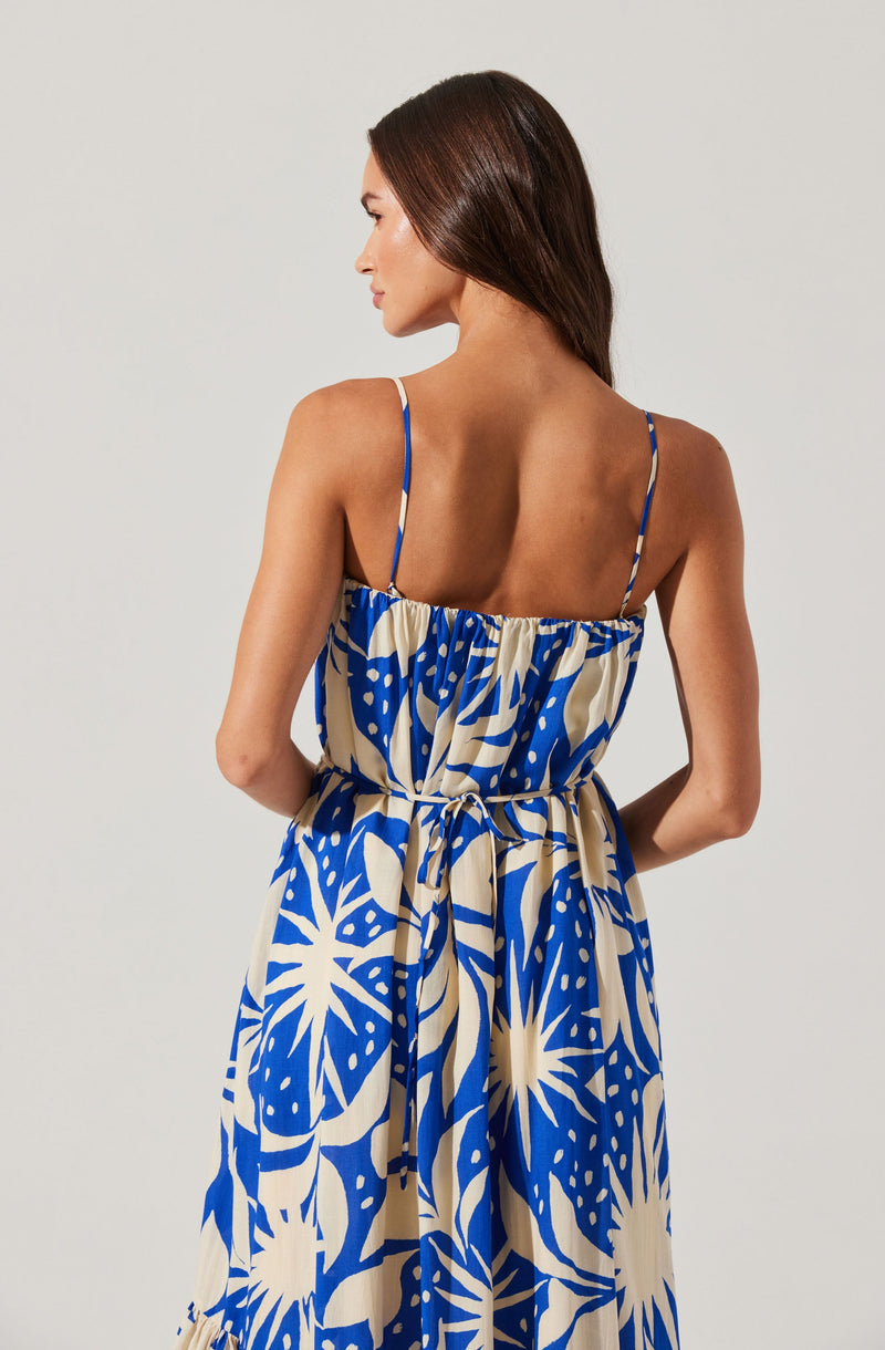 A woman is wearing a sleeveless dress with thin spaghetti straps. The dress features a bold, abstract pattern in blue and beige, with large leaf and sunburst shapes. The dress has a gathered waistline with a tie, creating a casual and flowing silhouette. The woman has long, brown hair and is posed with her back to the camera, standing against a plain, light-colored background.