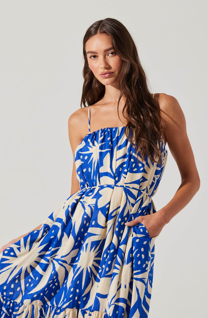 A woman wearing a sleeveless, blue and white patterned dress with thin spaghetti straps. The dress features a gathered neckline and a relaxed, flowy fit with pockets.