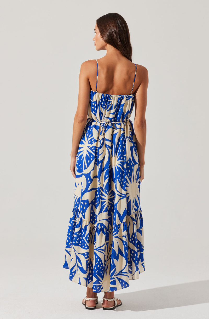 A woman is wearing a sleeveless, ankle-length summer dress with thin spaghetti straps. The dress features a vibrant, large floral and leaf pattern in blue, beige, and white colors. The fabric appears lightweight and flowy, suitable for warm weather.