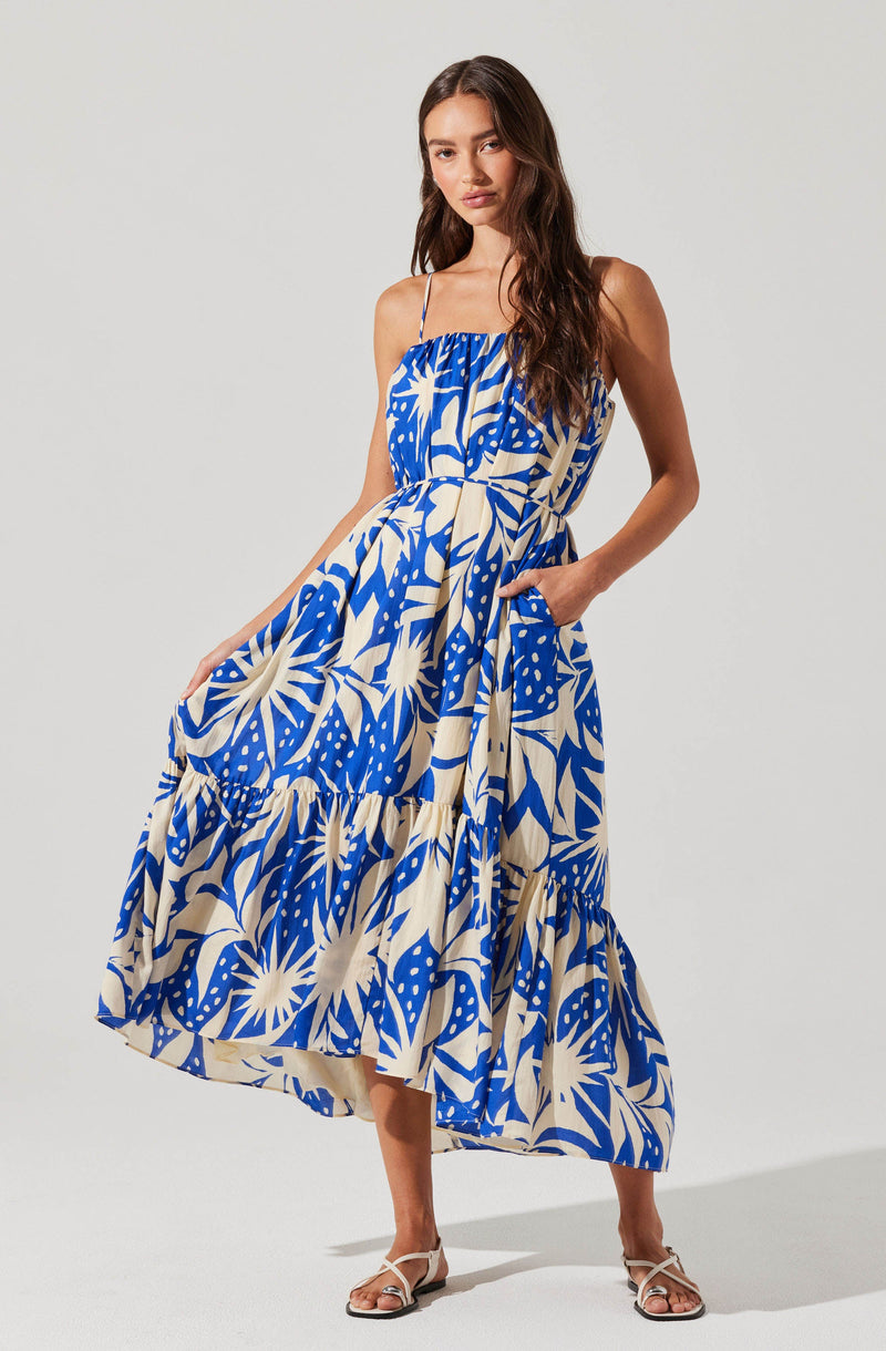 A woman is wearing a sleeveless, ankle-length dress with thin spaghetti straps. The dress features a bold, abstract floral pattern in shades of blue and off-white. The fabric appears lightweight, flowing, and gathered at the waist, creating a relaxed and elegant look. She has her right hand in a pocket and is wearing strappy white sandals.