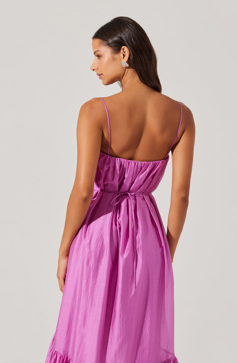 A woman is wearing a sleeveless, pink satin dress with thin spaghetti straps. The dress has a gathered bodice with a drawstring tie at the waist and a ruffled hem at the bottom. She is also wearing silver statement earrings.