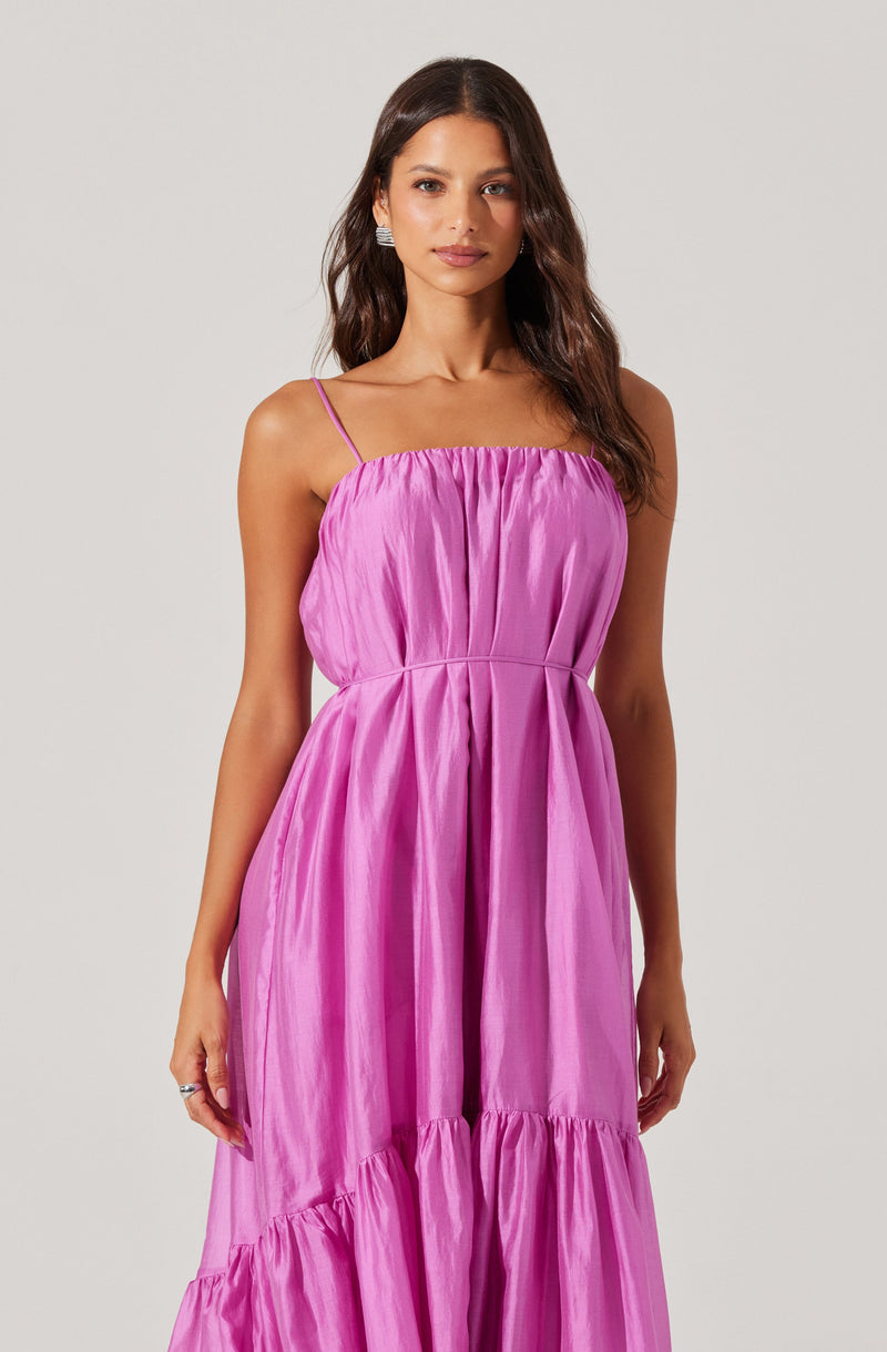 A woman is wearing a sleeveless, lavender-pink dress with thin spaghetti straps. The dress features a gathered, ruched bodice and a full-length skirt with gathered tiers. The fabric has a shiny, satin-like appearance.