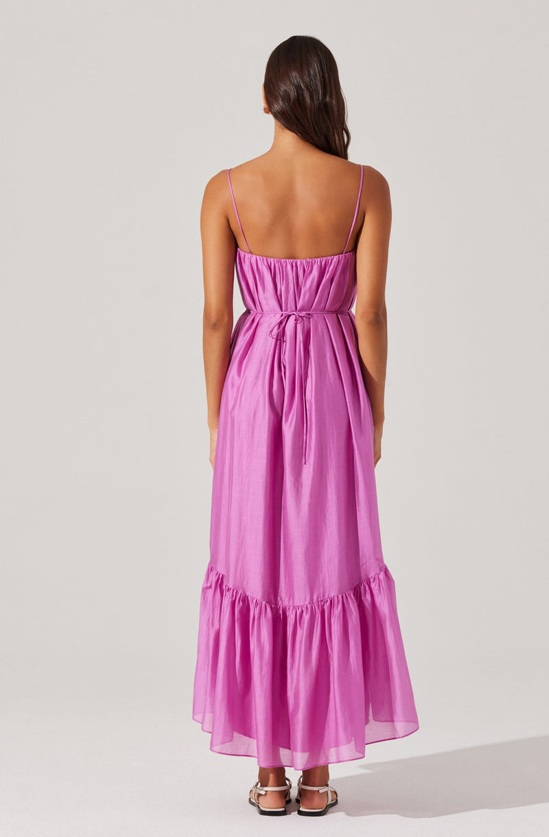 A woman is wearing a long, pink, sleeveless dress with thin shoulder straps. The dress has a gathered bodice and a slight cinch at the waist, with a fabric tie. The skirt is flowy and features a ruffled hem at the bottom.