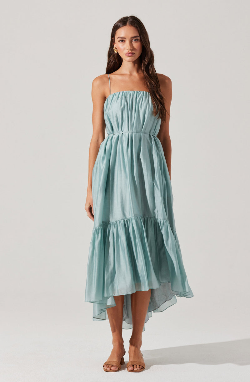 A woman is wearing a sleeveless, light blue maxi dress with thin spaghetti straps. The dress has a gathered, ruched bodice and a loose-fitting skirt with a ruffled hem that is slightly shorter in the front and longer in the back. She is also wearing open-toed, nude-colored high heels and gold hoop earrings.