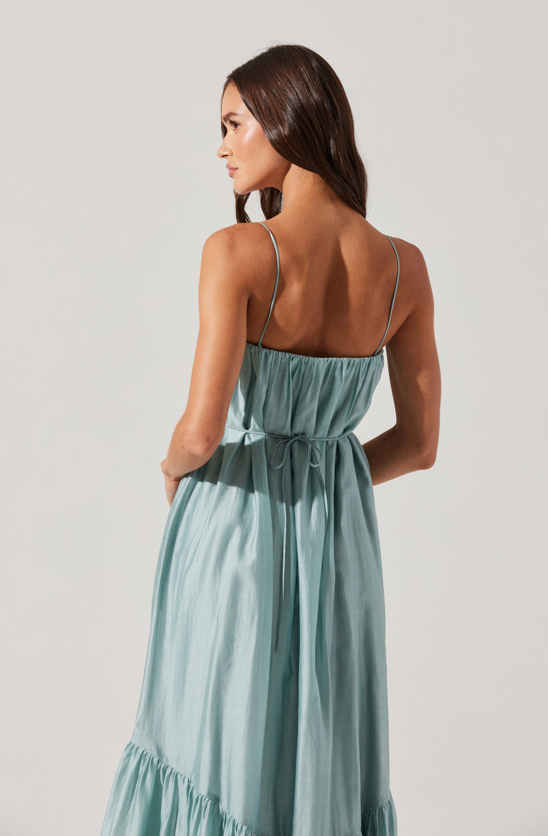 A woman is wearing a sleeveless, light blue satin dress with thin spaghetti straps. The dress has a gathered bodice and a waist tie that creates a cinched look. The skirt flows down with a ruffled hem at the bottom.