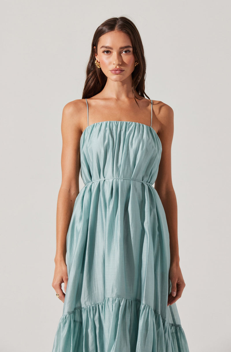 A woman is wearing a light blue, sleeveless dress with thin spaghetti straps. The dress features a gathered, ruched bodice and a flowing, tiered skirt with multiple pleated sections. The fabric appears lightweight and shiny, creating a soft, elegant look.