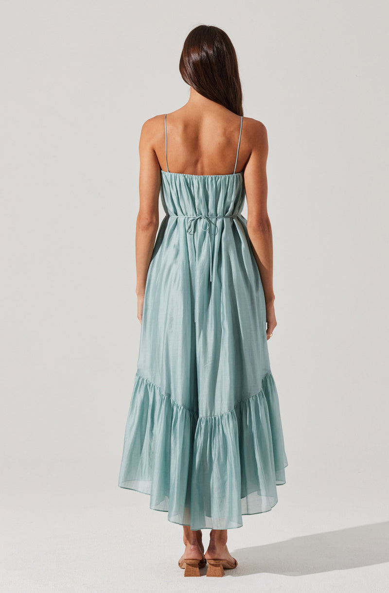 A woman is wearing a sleeveless, light blue, flowy dress with spaghetti straps. The dress features an elastic bodice and a tie detail at the waist, with a tiered, ruffled hem that reaches down to her ankles. She is also wearing open-toe, block-heeled shoes.