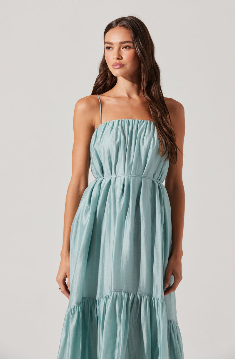 A woman is wearing a sleeveless, light blue dress with spaghetti straps. The dress features a gathered, ruched bodice and a waistline seam, with a flowing skirt that has a tiered design with gathered fabric.