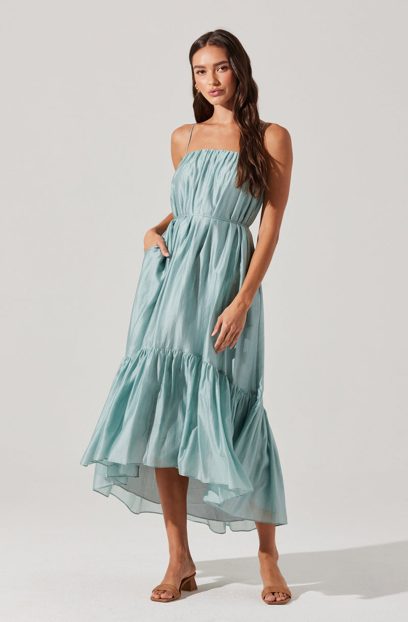 A woman is wearing a light blue, sleeveless, maxi dress with thin spaghetti straps. The dress has a gathered, ruched bodice and a tiered, ruffled hemline that flows to her ankles. She is also wearing tan open-toe heels.