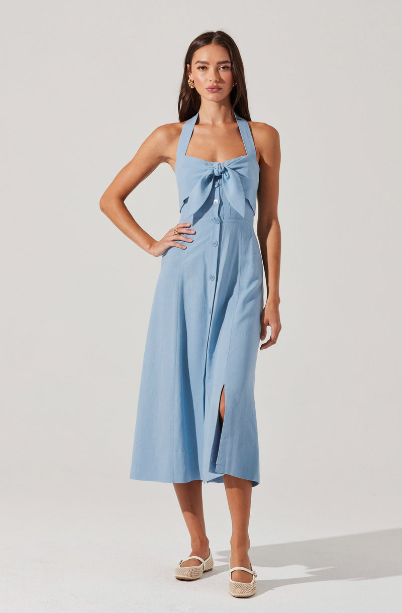A woman is wearing a light blue sleeveless dress with wide straps that form a halter neckline. The dress features a large bow detail on the chest, a button-down front, and a slit in the skirt. She is also wearing white, woven, closed-toe shoes.