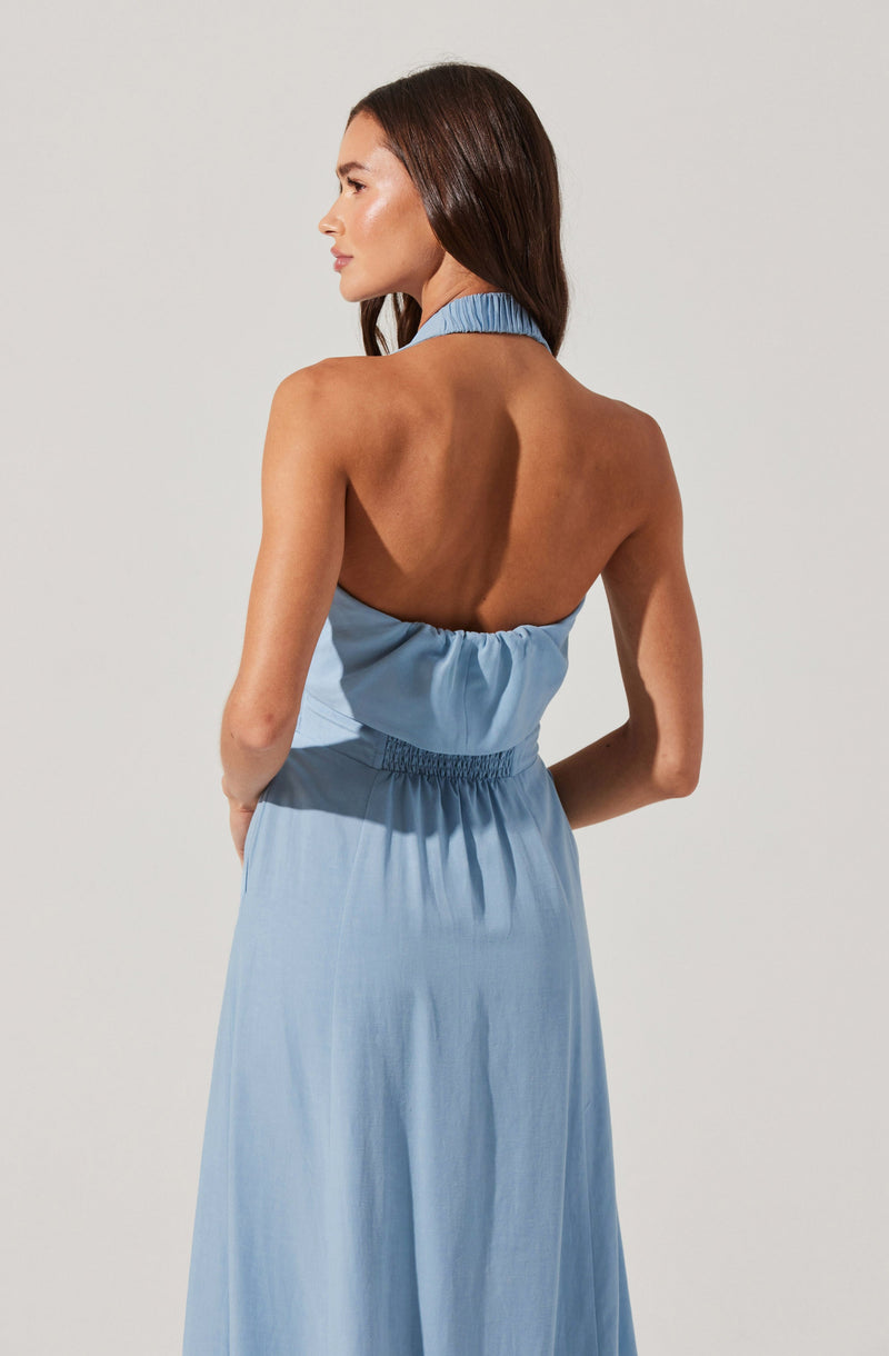 A woman is wearing a light blue, sleeveless dress. The dress has a high, ruffled collar around the neck and an open back with gathered fabric below the neckline. The waist is cinched with elastic, and the skirt is long and flowing, made of a lightweight fabric.