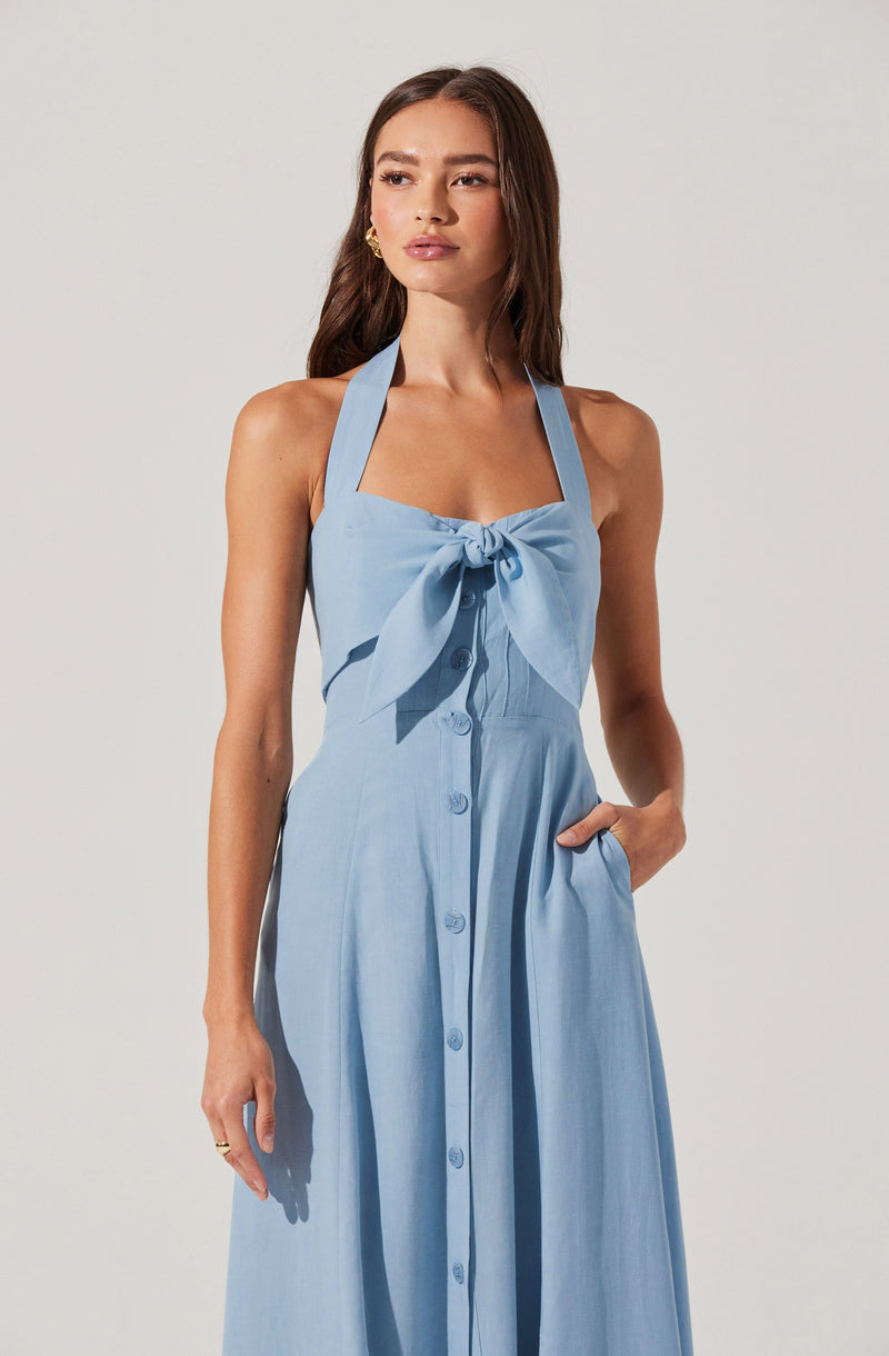 A woman is wearing a sleeveless, light blue dress with wide shoulder straps that form a halter neckline. The dress features a large bow at the chest and is buttoned down the front with matching blue buttons. She has one hand in her pocket and is accessorized with small gold earrings.